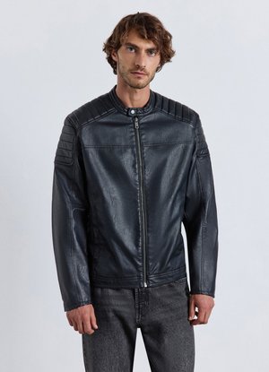 Young man with curly hair wearing a black zippered leather jacket and dark jeans standing against a plain light background.