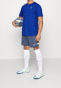 Modrá vzorovaná atletická košile s krátkým rukávem, šedé šortky, bílé ponožky a bílé boty s modrými akcenty. Drží šedý fotbal Nike.