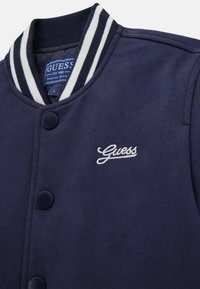 Giacca bomber blu navy con colletto a coste a righe, bottoni a pressione e logo "Guess" ricamato bianco sul petto. Tessuto morbido, design casual.