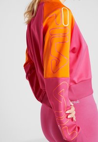 Kvinna iklädd en rosa och orange FILA-tröja och rosa leggings, med FILA-logotyp tryckt på ärmen och midjebandet.