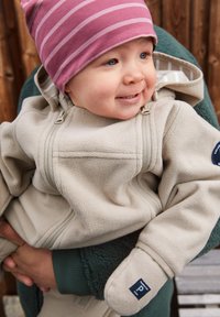 Beige Fleece-Jacke mit einem Reißverschluss vorne, ausgestattet mit Kapuze und Handschuhen, getragen von einem Kind. Eine rosa gestreifte Mütze ergänzt das Outfit.