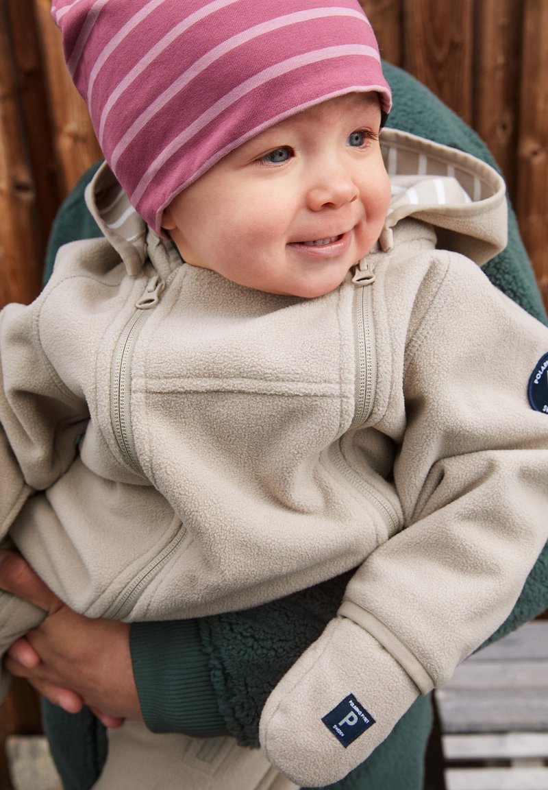 Beige Fleece-Jacke mit einem Reißverschluss vorne, ausgestattet mit Kapuze und Handschuhen, getragen von einem Kind. Eine rosa gestreifte Mütze ergänzt das Outfit.