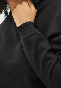 Schwarzer Sweatshirt aus glattem Stoff, mit einem gerippten Bündchen und einem gestickten Sternenmotiv am Ärmel. Nahaufnahme des Arms.