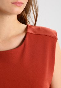 Rdzawy bezrękawnik z szerokim dekoltem i teksturowaną tkaniną, z gładką linią ramion i minimalistycznym designem.