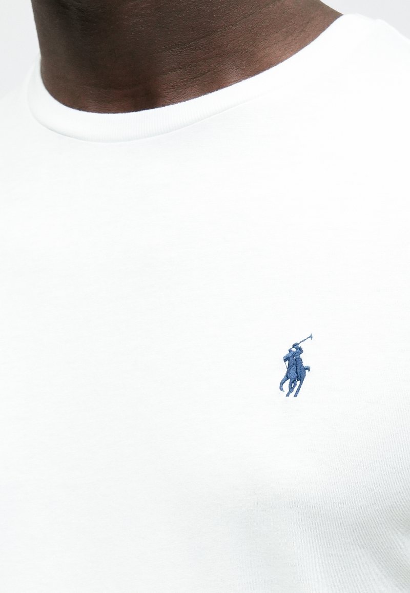 Maglietta bianca in cotone con un piccolo logo ricamato di un giocatore di polo blu navy sul petto a sinistra. Collo rotondo e tessuto liscio.