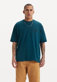 Tricou din bumbac teal cu mâneci scurte, prezentând un logo subtil negru Levi's pe piept. Asortat cu pantaloni bej, purtat cu un lanț argintiu.