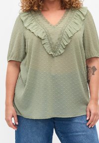 Blusa verde chiaro con scollatura a V, caratterizzata da un bordo arricciato e una texture a pois. Maniche corte e vestibilità ampia.