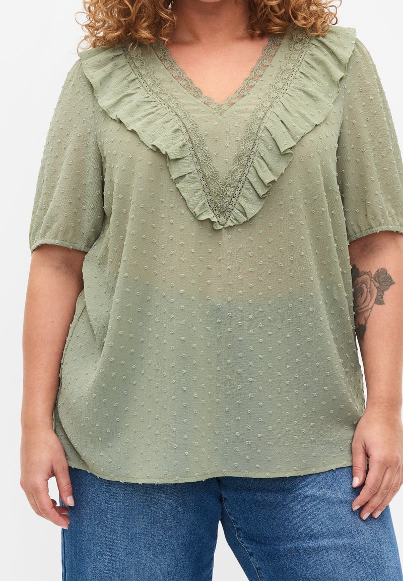 Blusa verde chiaro con scollatura a V, caratterizzata da un bordo arricciato e una texture a pois. Maniche corte e vestibilità ampia.