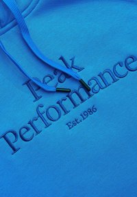 Niebieska tkanina bluzy z kapturem z niebieskimi sznurkami, z wyhaftowanym napisem "Peak Performance Est. 1986" w dopasowanym niebieskim kolorze nici.