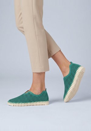 Persoon die turquoise instappers draagt met een getextureerd patroon en crèmekleurige zolen, gecombineerd met beige cropped broeken tegen een effen achtergrond.