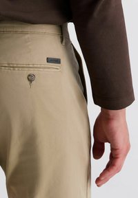 Beige Hosen aus glattem Stoff, ausgestattet mit einer Rückentasche mit Knopf und einem kleinen Logo-Tag. Braunes Langarmhemd ist sichtbar.
