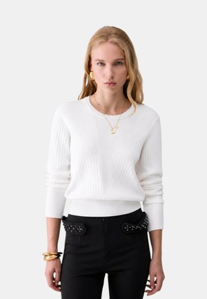 Donna bionda che indossa un maglione bianco a coste, pantaloni neri con tasche borchiate, orecchini d'oro, collana e braccialetti, in piedi su sfondo bianco.