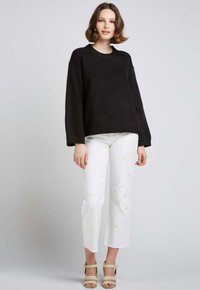 Pull noir à encolure ronde et manches longues, associé à un pantalon blanc court avec broderies florales jaunes et des sandales beiges.