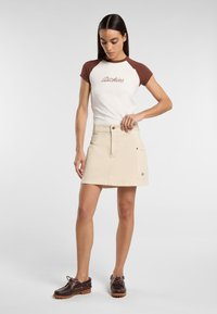 Beige denimkjol med en framficka och logotypapplikation, tillsammans med en vit och brun raglantröja och bruna snörskor.