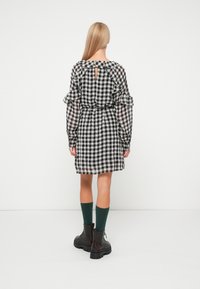 Robe à carreaux noir et blanc avec des manches à volants, un dos en forme de trou de serrure, une taille cintrée, et accompagnée de chaussettes vertes et de bottes militaires noires.
