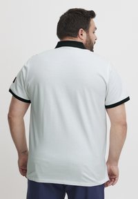 Bijela pamučna polo majica s crnim ovratnikom i obrubima rukava, s glatkom teksturom i malom smeškastom etiketom na lijevom ramenu.