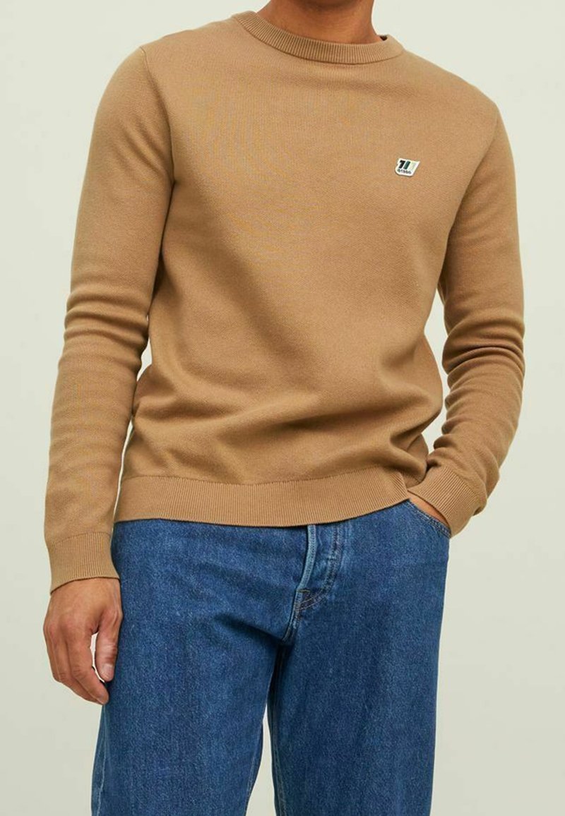 Vaaleanruskea joustinneuleinen puuvillaneule, jossa pieni logo-ompelu rintakehässä. Yhdistetty sinisiin farkkuihin vaaleaa taustaa vasten.