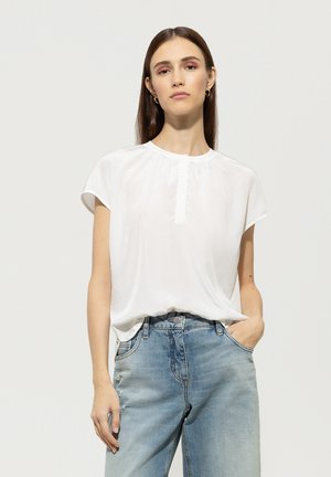 Weiße Bluse mit kurzen Ärmeln, rundem Ausschnitt und kleiner Knopfleiste vorne. Kombiniert mit hellblauen Jeans. Glatte Stoffoberfläche.
