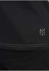 Camiseta negra de algodón con un pequeño logotipo bordado en blanco con flechas en el dobladillo, y costuras blancas contrastantes a lo largo de la parte inferior.