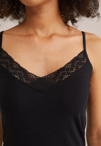 Nahaufnahme einer Frau, die ein schwarzes Spaghettiträgershirt mit dünnen Trägern und floraler Spitzenverzierung entlang des V-Ausschnitts trägt.