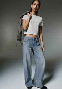 Vit kortärmad t-shirt med text, ljusblå vida jeans och svarta klackstövlar. Modellen håller en grå jacka över axeln.