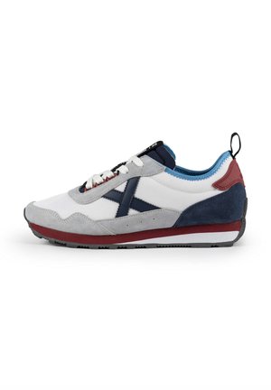 Zapatilla blanca y gris con acentos en azul marino, rojo y azul. Materiales de ante texturizado y malla. Diseño con cordones y un logotipo prominente en el lateral.