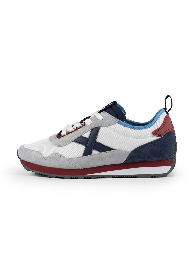 Zapatilla blanca y gris con acentos en azul marino, rojo y azul. Materiales de ante texturizado y malla. Diseño con cordones y un logotipo prominente en el lateral.