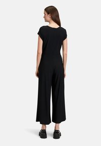 Zwarte jumpsuit met een aansluitend lijfje, korte mouwen en wijde benen. Kenmerkt zich door een ritssluiting aan de achterkant en een gladde stofstructuur.