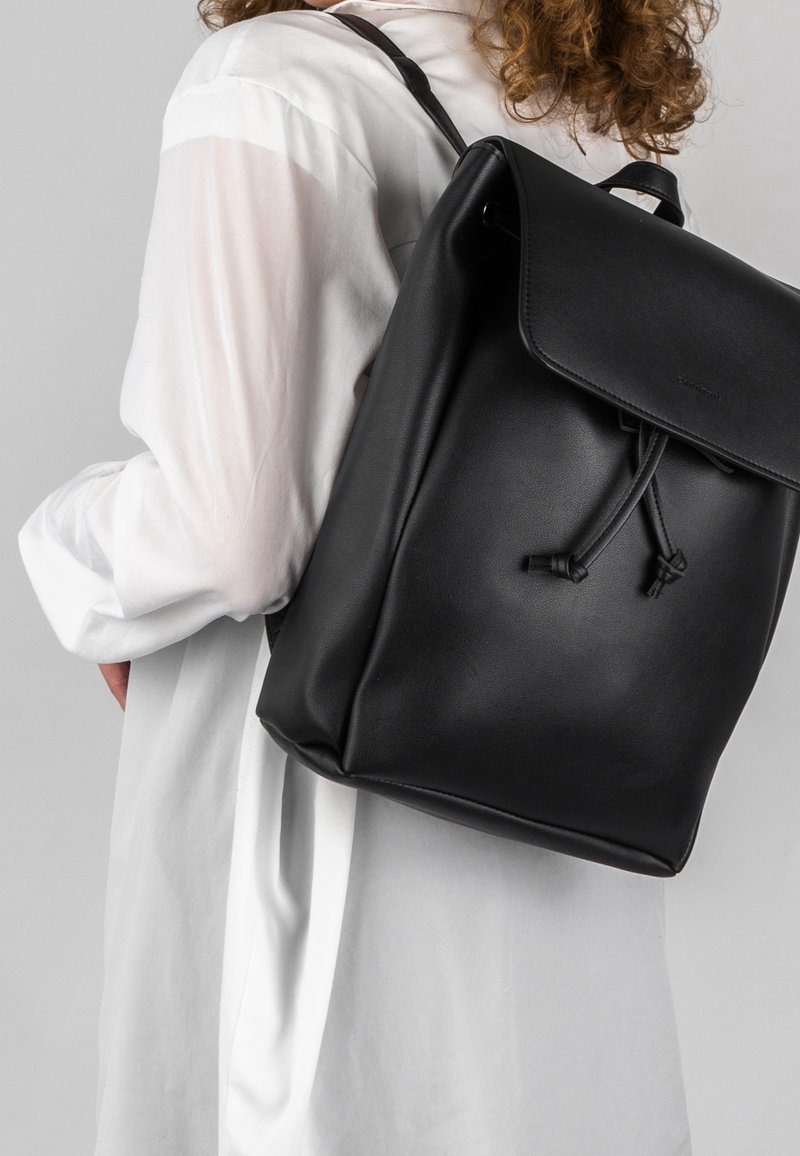 Schwarzer Leder-Rucksack mit Klappenverschluss, verstellbaren Trägern und Kordelzugsdetail; vor einem hellgrauen Hintergrund präsentiert.