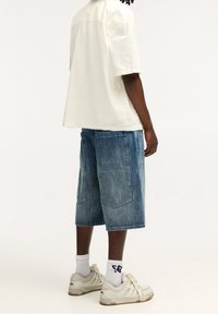 Loszittend crèmekleurig overhemd, denim short met vervaagde textuur en zijzakken, witte sneakers met tonale accenten en geribbelde sokken.