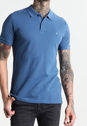 Polo de algodón azul con cuello clásico, mangas cortas y un logotipo sutil en el pecho. Combinado con jeans oscuros.