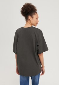 Donna con capelli ricci raccolti, indossa una maglietta a maniche corte grigio scuro e jeans blu, rivolta di spalle su uno sfondo semplice.