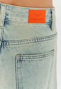 Světle modré džíny z denimu s texturovaným povrchem, vybavené oranžovou etiketou s nápisem "vytvořte se" poblíž pasu pro branding.