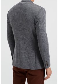 Grå rutmönstrad blazer med en struktur i tyget, slagkrage och fyra bruna knappar på ärmarna. Bruna byxor synliga längst ner.