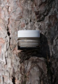 Korres Bounce Firming Intense Moisturizer i en genomskinlig burk med ett vitt lock, med brun och svart märkning och text på en texturerad yta.