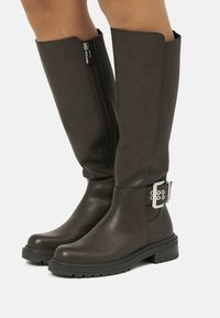 Knöchelhoche, dunkelbraune Lederstiefel mit seitlichem Reißverschluss und Schnallendetails. Robuste Gummisohle mit strukturierter Oberfläche.