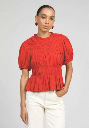 Femme aux cheveux plaqués en arrière porte un chemisier rouge texturé à manches ballon et un pantalon blanc, posant avec une main dans la poche sur un fond clair.