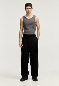 Un débardeur gris sans manches associé à un pantalon large taille haute noir, avec une fermeture à boutons. Le mannequin se tient les mains dans les poches.