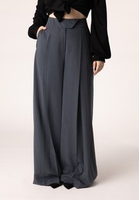 Brede, geplooide broeken van donkergrijze stof met een hoge taille en zijzakken, gecombineerd met een zwart top en zwarte schoenen.