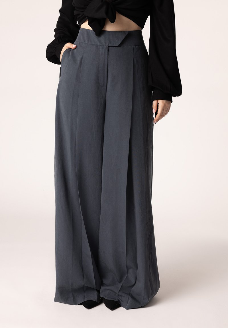 Brede, geplooide broeken van donkergrijze stof met een hoge taille en zijzakken, gecombineerd met een zwart top en zwarte schoenen.