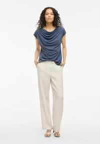 Mörkblå draperad topp med en mjuk textur, kombinerad med beige linnebyxor med vida ben. Modellen står med händerna i fickorna och har sandaler på sig.