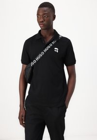 Μαύρο πόλο μπλουζάκι με κλασικό γιακά, που διαθέτει ένα μικρό λευκό λογότυπο στο στήθος. Φοράει με σκούρους παντελόνες και μια μαύρη τσάντα ώμου.