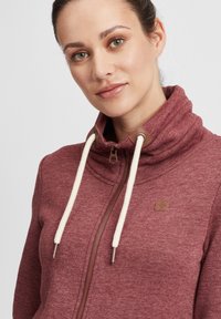 Sweatshirt zippé bordeaux avec col haut, cordons de tirage crème et accents en cuir marron. Texture douce et léger motif mélangé visible.