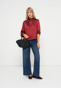 Rote Satinbluse mit Stehkragen, kombiniert mit weit geschnittenen Jeans und einer schwarzen Handtasche. Das Model steht vor einem schlichten Hintergrund.