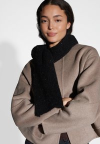 Sciarpa nera texturizzata che si drappeggia intorno a un cappotto oversize color marrone chiaro con vestibilità rilassata, caratterizzato da tessuto liscio e dettagli sottili.