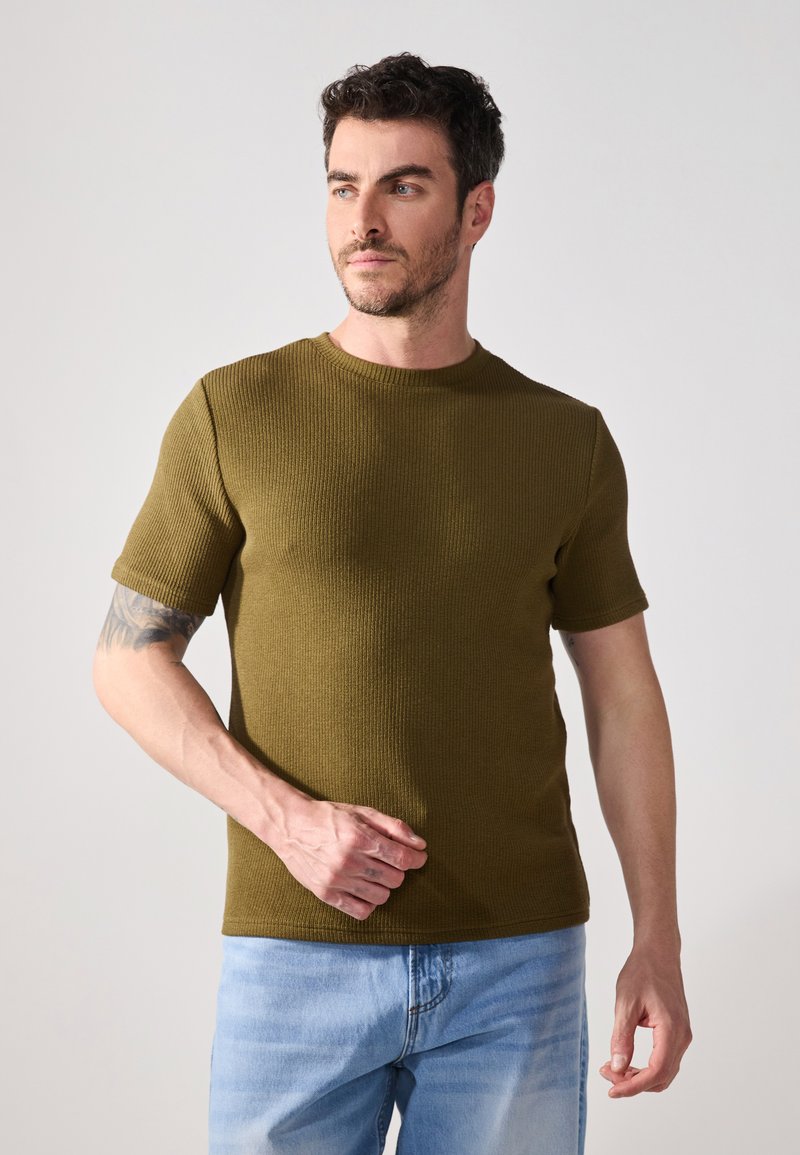 Tricou scurt cu mânecă scurtă de culoare verde măsliniu, cu guler rotund, completat cu blugi din denim de culoare albastru deschis. Detalii de țesătură sunt vizibile.