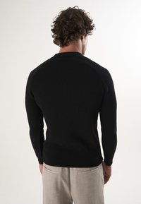 Must jaotatud kudum, millel on pikad varrukad, ümmargune kaelus ja keha lähedane siluett. Kantud heledate hallide pükstega.