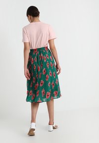 Gonna verdi plissettata con motivo floreale rosso, abbinata a una t-shirt rosa chiaro. Presenta una cintura elastica e sandali con tacco a blocco bianchi.