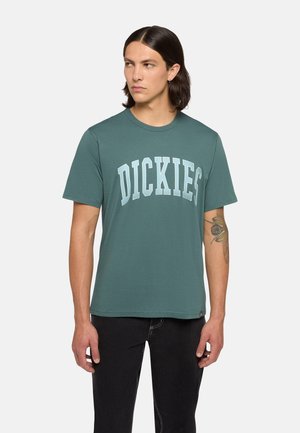 Teal kortærmet t-shirt med runde halsudskæring og "DICKIES" trykt i lyseblå, lavet af blødt bomuldsblandingsmateriale, tætsiddende design, sammen med sorte jeans.