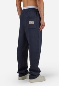 Nachtblaue Baumwollmischung-Loungehosen mit gerippter Textur, elastischem Bund und Gesäßtasche mit eingewebtem Etikett. Kombiniert mit weißen Sneakern.